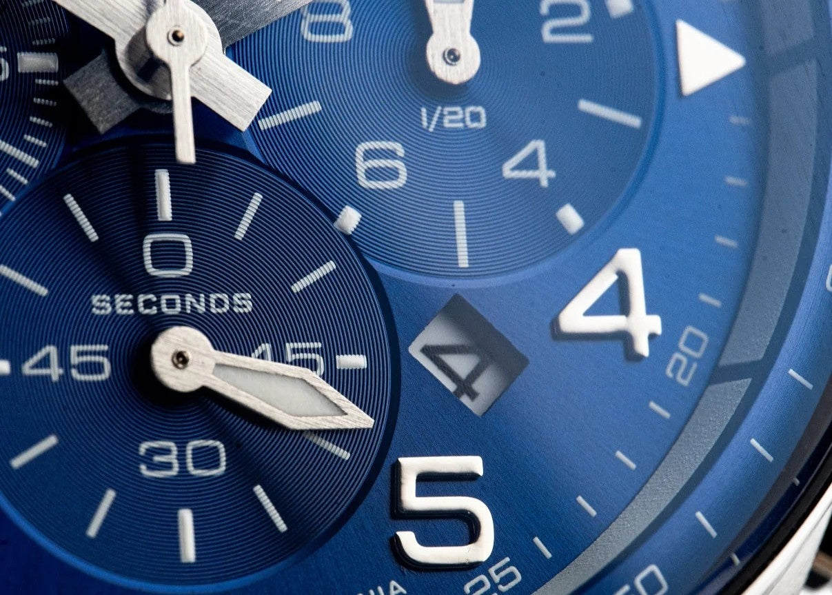 Macro view of a blue Vostok Europe Space Race chronograph dial showing the timing subdials, date window, luminous silver hands, raised numerals and detailed racing-inspired design